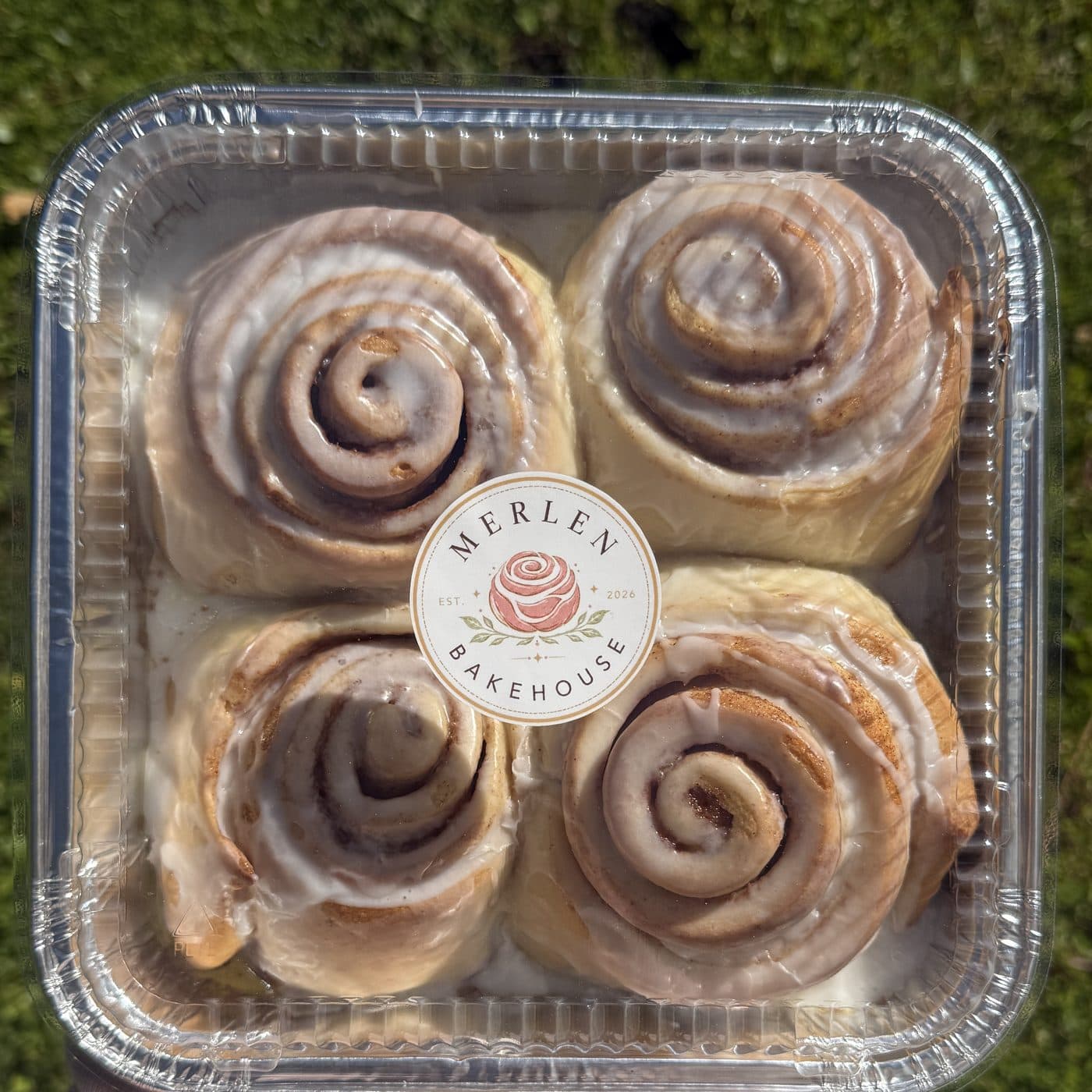 A box of four Merlen Bakehouse cinnamon rolls with the rose sticker, held over grass